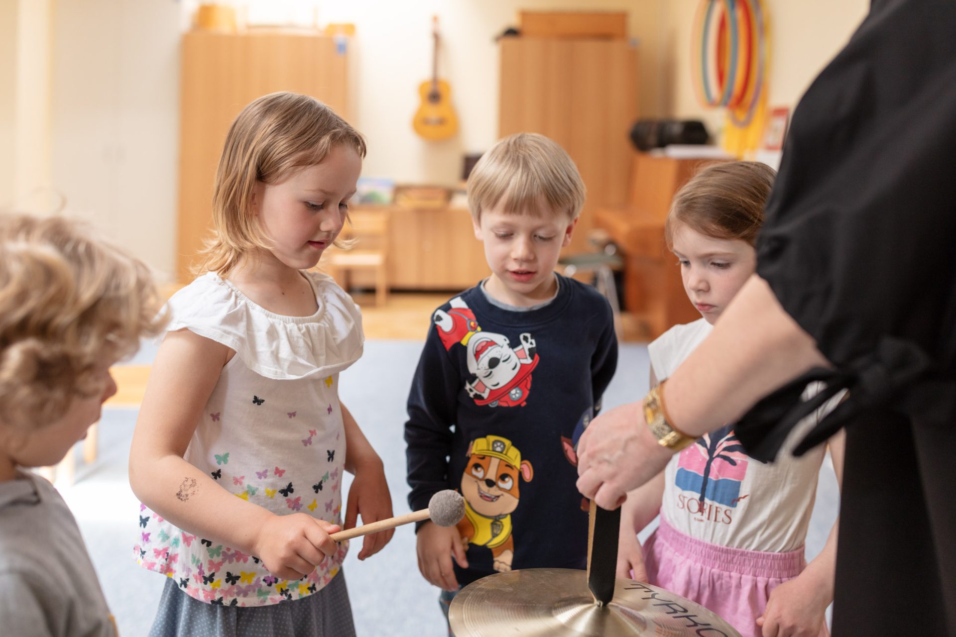 vier Kinder, die im Kreis stehen, jemand hält ein Becken in die Mitte, ein Kind hält einen Stick in der Hand