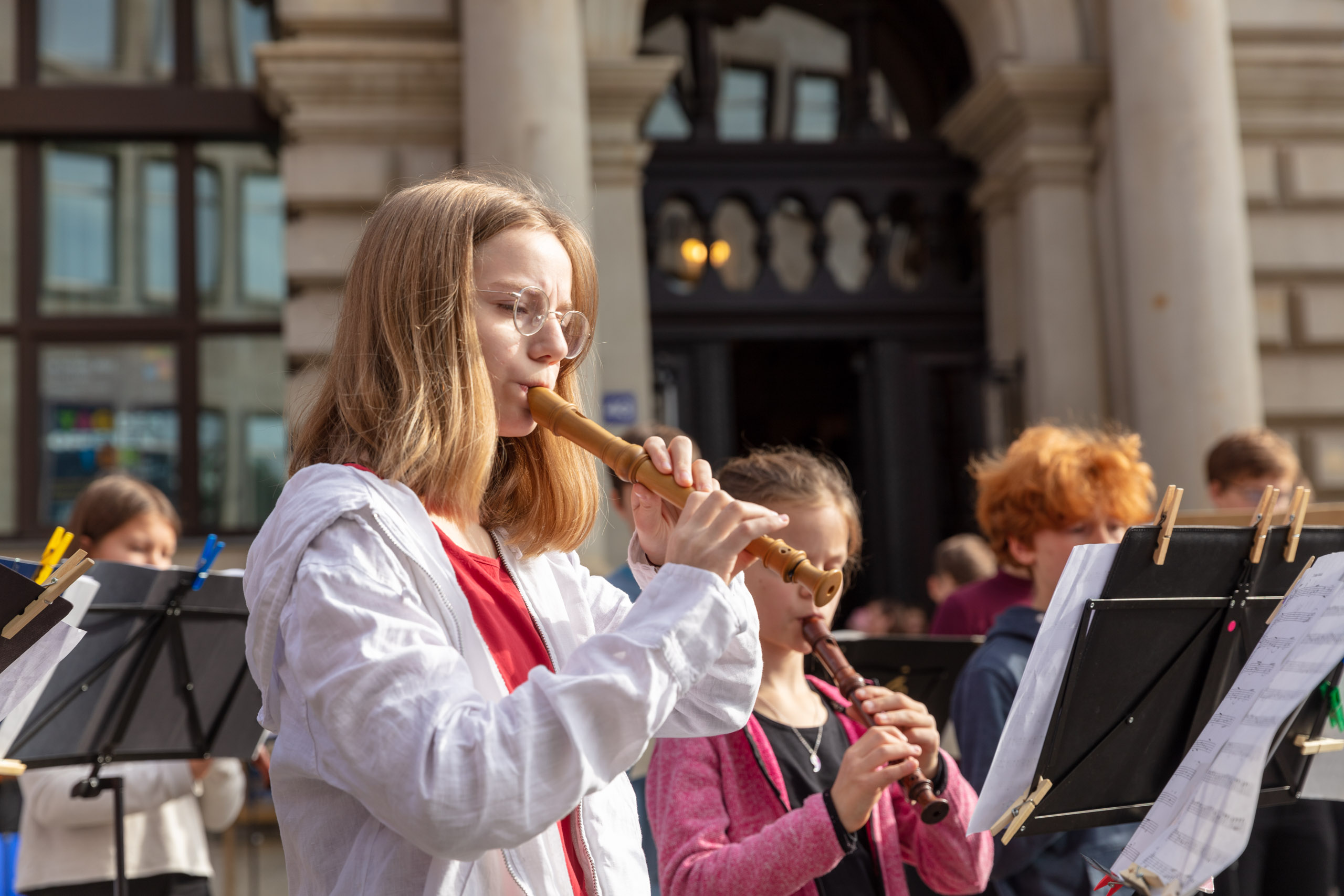 eine Gruppe von Schüler:innen musiziert vor der Musikschule, im Vordergrund spielt ein Mädchen Blockflöte