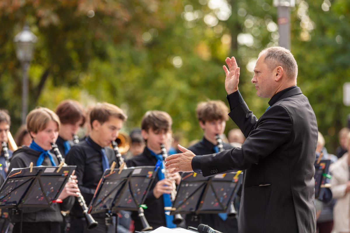 das Sinfonische Blasorchester spielt vor der Musikschule, im Vordergrund steht der Dirigent Wilfried Thoß, im Hintergrund spielt eine Gruppe von Schüler:innen Klarinette