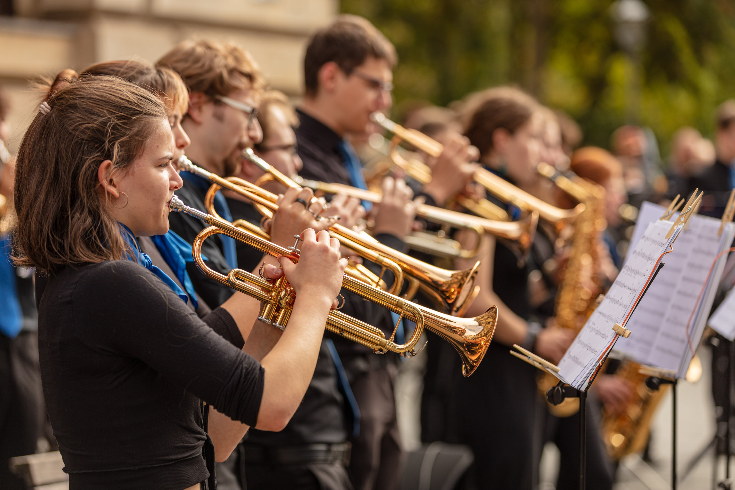 Mitglieder des Sinfonischen Blasorchesters spielen draußen vor der Musikschule, im vordergrund spielt ein Mädchen mit blauem Tuch Trompete, im Hintergrund spielen andere Schüler:innen ebenfalls Trompete und Saxophon