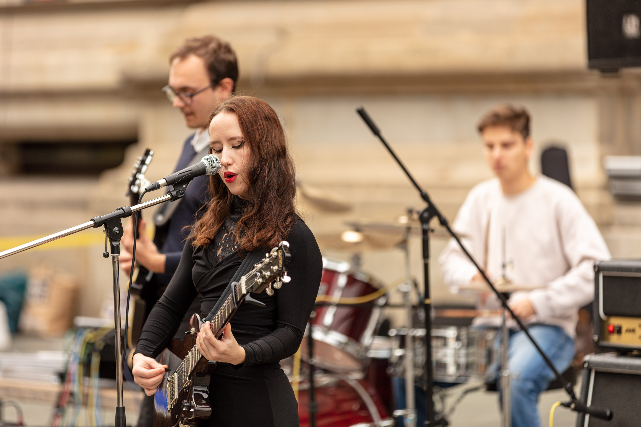 drei Schüler:innen spielen draußen vor der Musikschule, im Vordergrund steht ein Mädchen, das Gitarre spielt und singt, hinter ihr spielt ein Junge Gitarre und im Hintergrund spielt ein Junge Schlagzeug