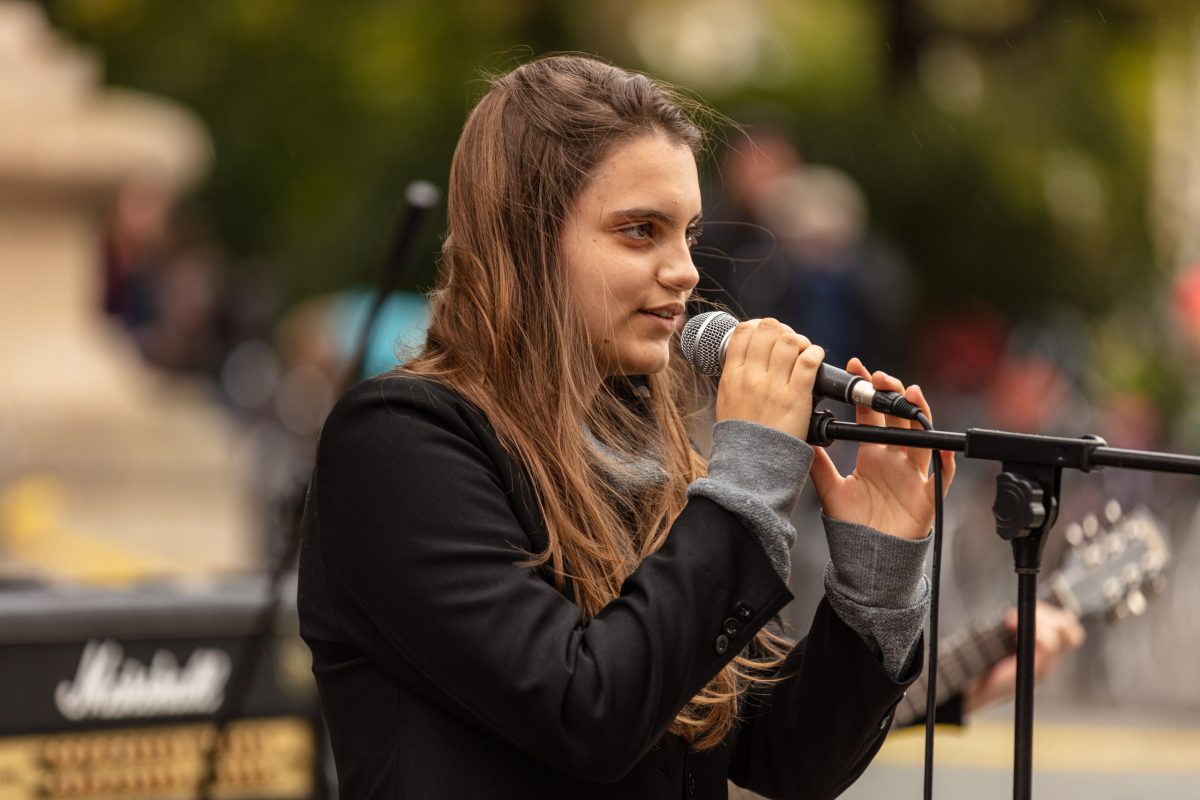 ein Mädchen hält ein Mikrofon und singt, im Hintergrund spielt jemand Gitarre