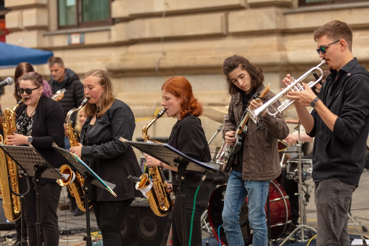 eine Gruppe SChüler:innen spielt vor der Musikschule, drei Schülerinnen spielen links Saxophon, daneben spielt ein Schüler Gitarre, ganz recehts spielt ein Schüler Posaune