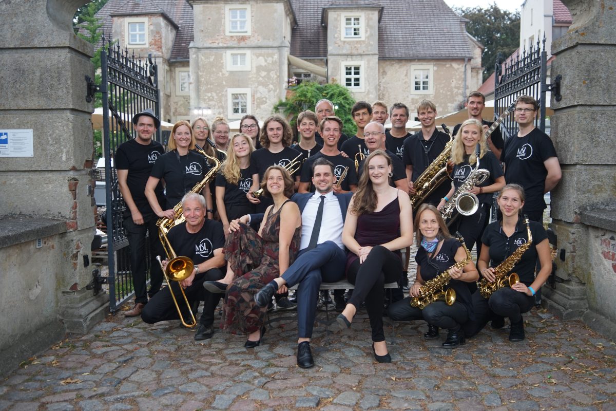 Mitglieder der MSL Bigband stehen in einem großen historischen Hoftor, viele tragen ein schwarzes Shirt mit dem Bigband Logo, die Musiker:innen halten ihre Instrumente, darunter Saxophon, Posaune und Gitarre