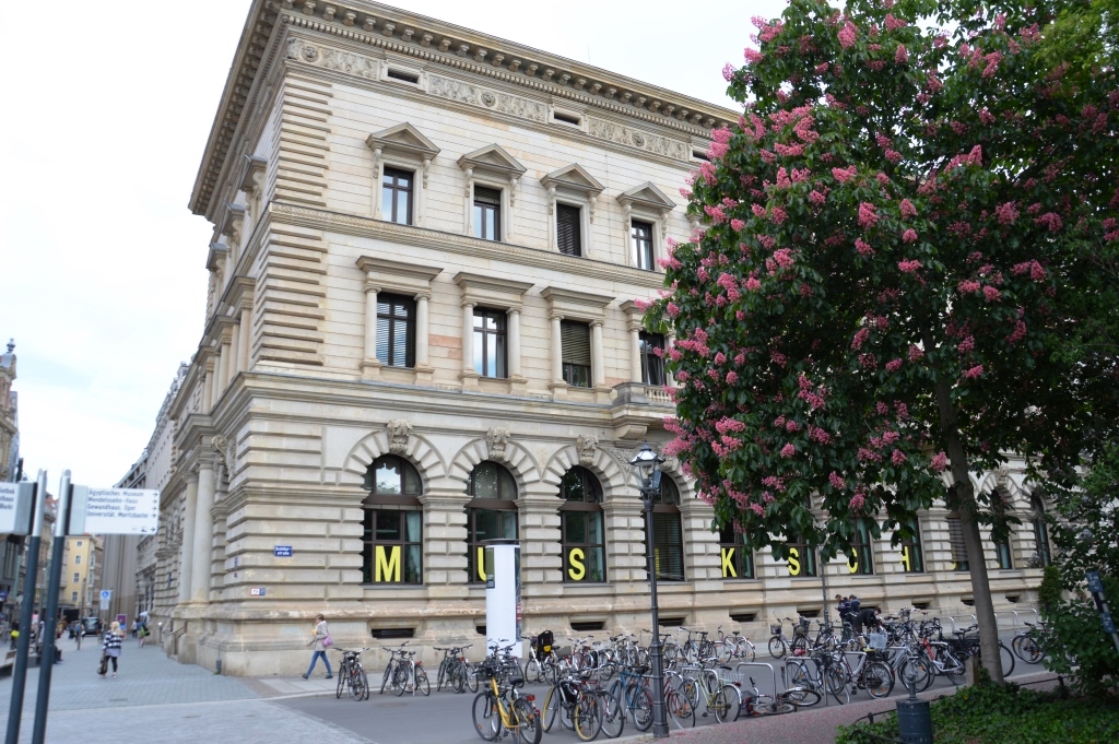 Hauptstelle, Petersstraße, ein dreistöckiges Gebäude im Neorenaissance-Stil mit aufwendig verzierter Fassade, im Vordergrund steht ein Baum mit hellrosaBlüten