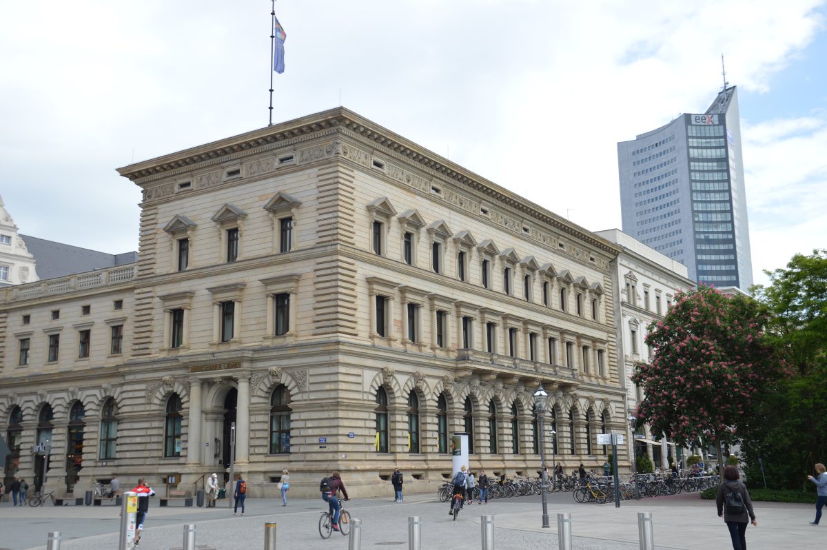 Vorderansicht der Hauptstelle, Petersstraße, ein dreistöckiges Gebäude im Neorenaissance-Stil mit aufwendig verzierter Fassade und zwei großenSäulen am Eingang, im Hintergrund sieht man das City-Hochhaus