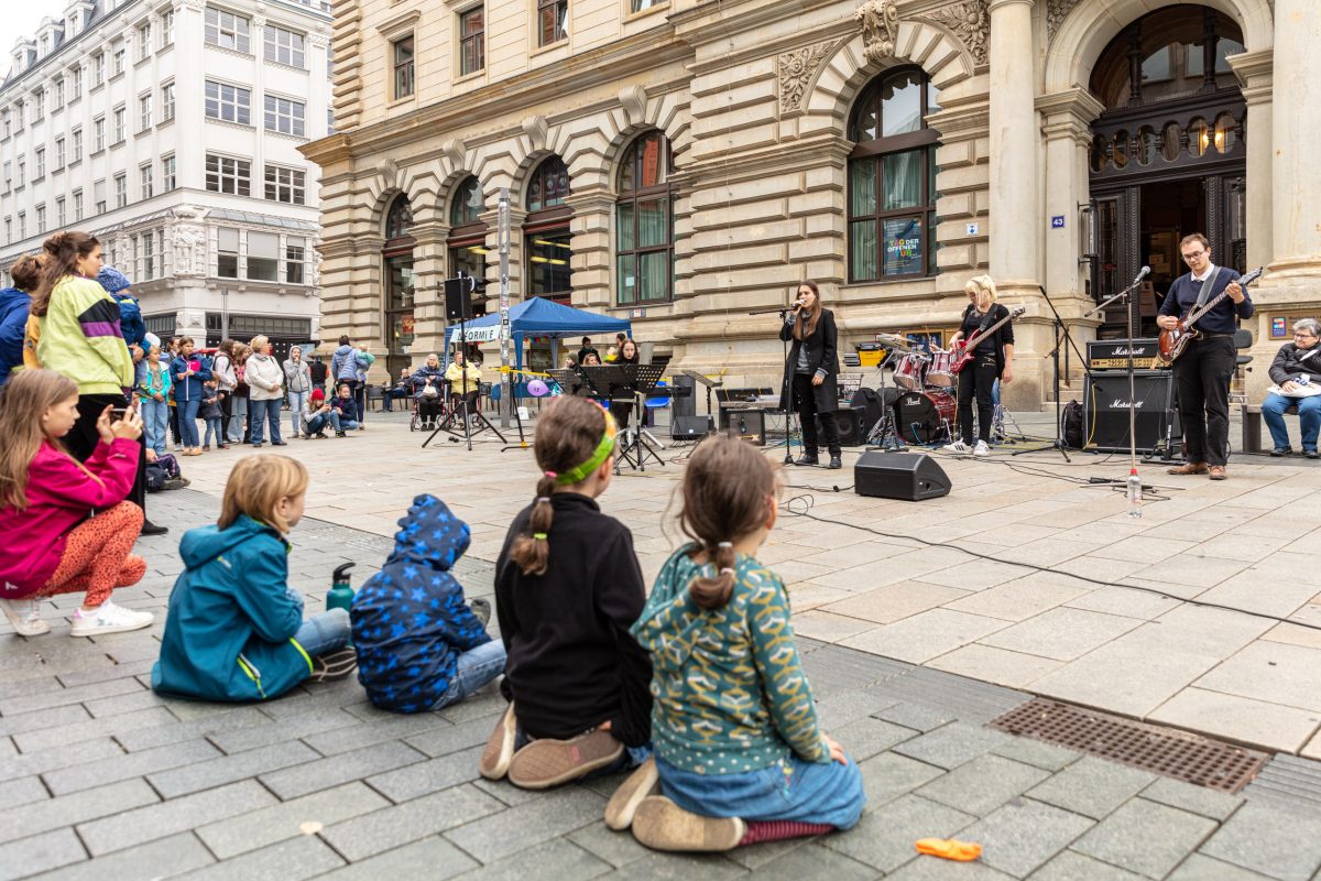 Kinder sitzen beim Tag der offenen Tür vor der Musikschule und hören einem Ensemble zu