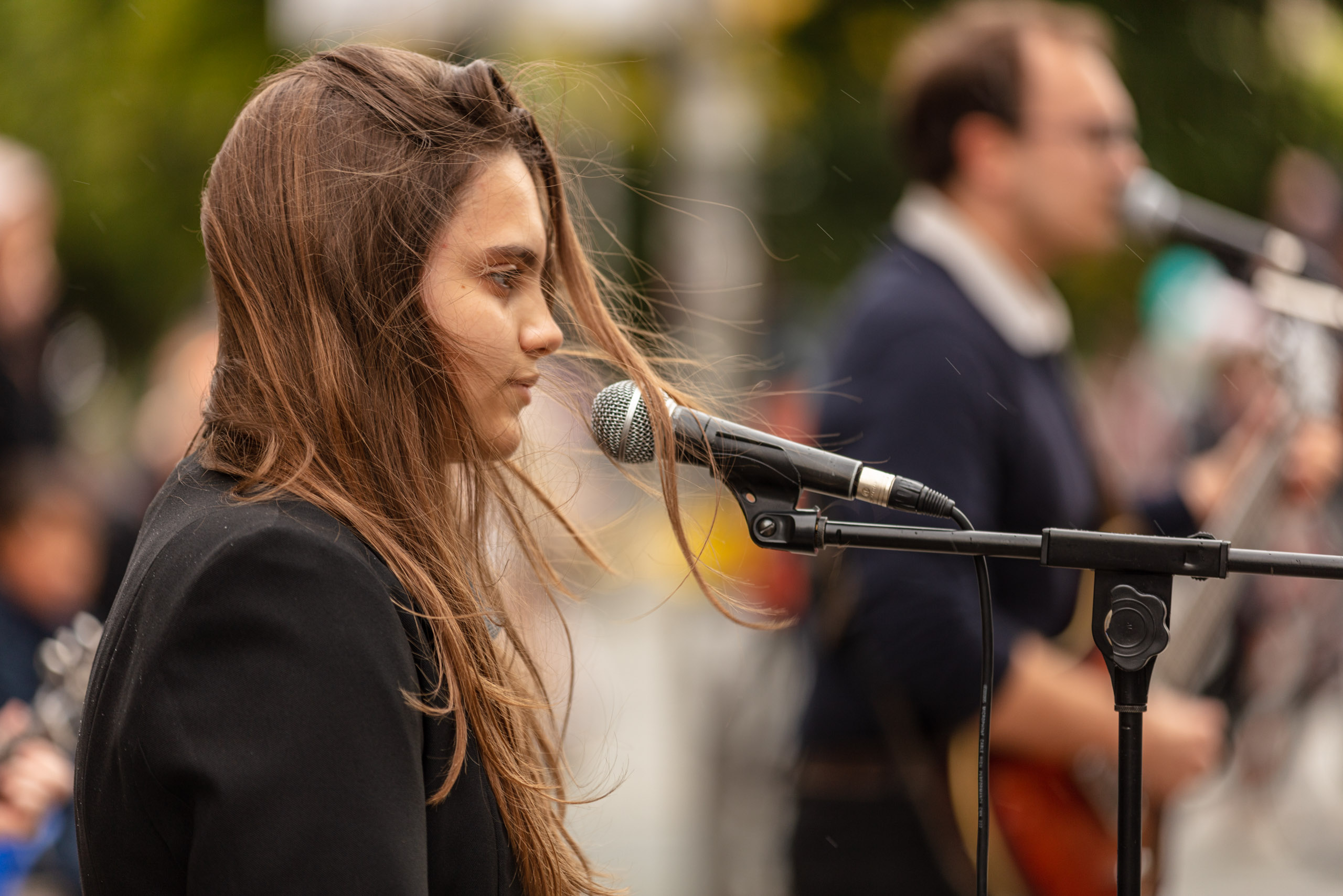 eine junge Frau steht an einem Mikrofon, ihre Haare wehen im Wind, im Hintergrund singt ein Mann und spielt Gitarre