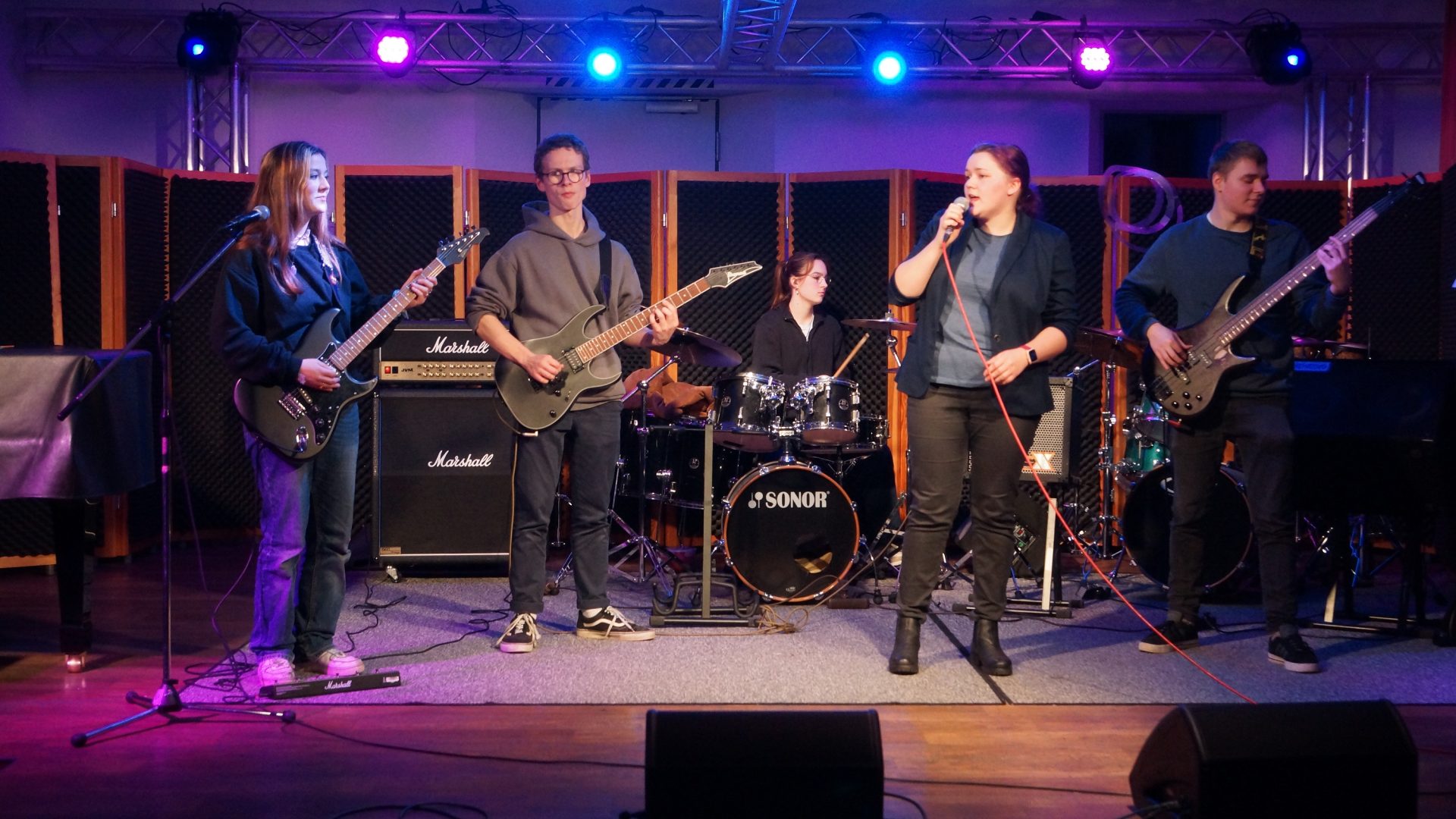 die Billie Peppers treten auf einer blau - violett beleuchteten Bühne auf, links zwei Gitarrist:innen, mittig die Schlagzeugerin und Sängerin und rechts der Bassist